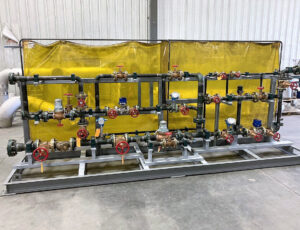 Custom carbon steel piping skid made exactly to client specs, featuring valves, controls, gauges, bypasses, and filters. Shown with yello backdrop and stainless steel frame