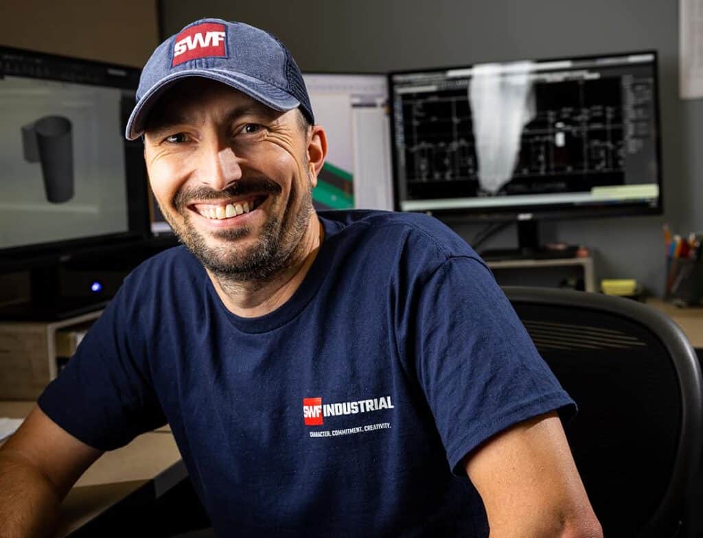SWF team member smiling in a blue uniform shirt and baseball cap with red letters SWF in front of computer TEKLA 3d modeling software used for steel projects