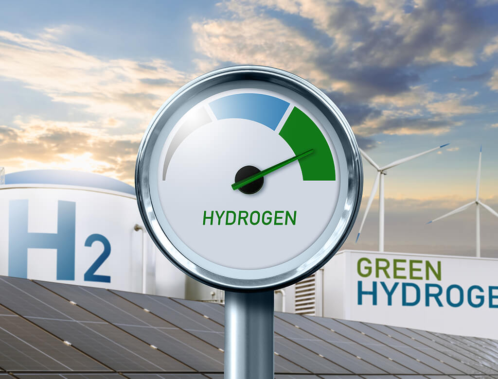 A dial with the hand reading "green," and the word HYDROGEN below, in front of white buildings with text reading H2 and Green Hydrogen.