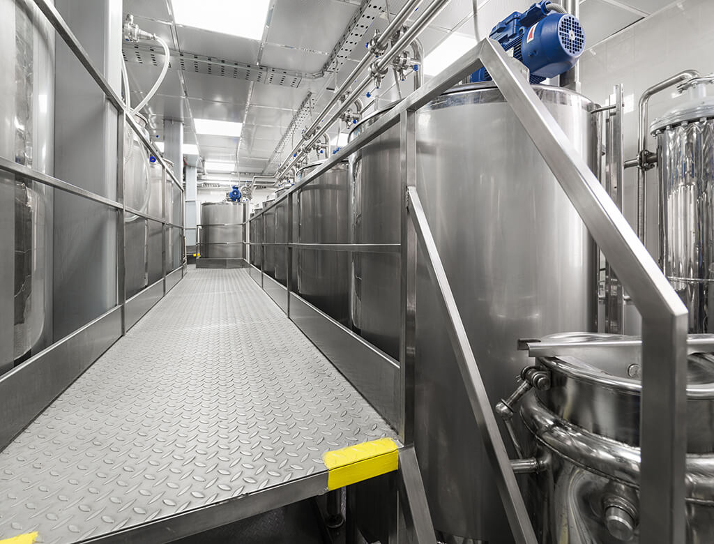 Stainless steel infrastructure for pharmaceutical plant showing OSHA railings and stairs on a catwalk between large tanks. 