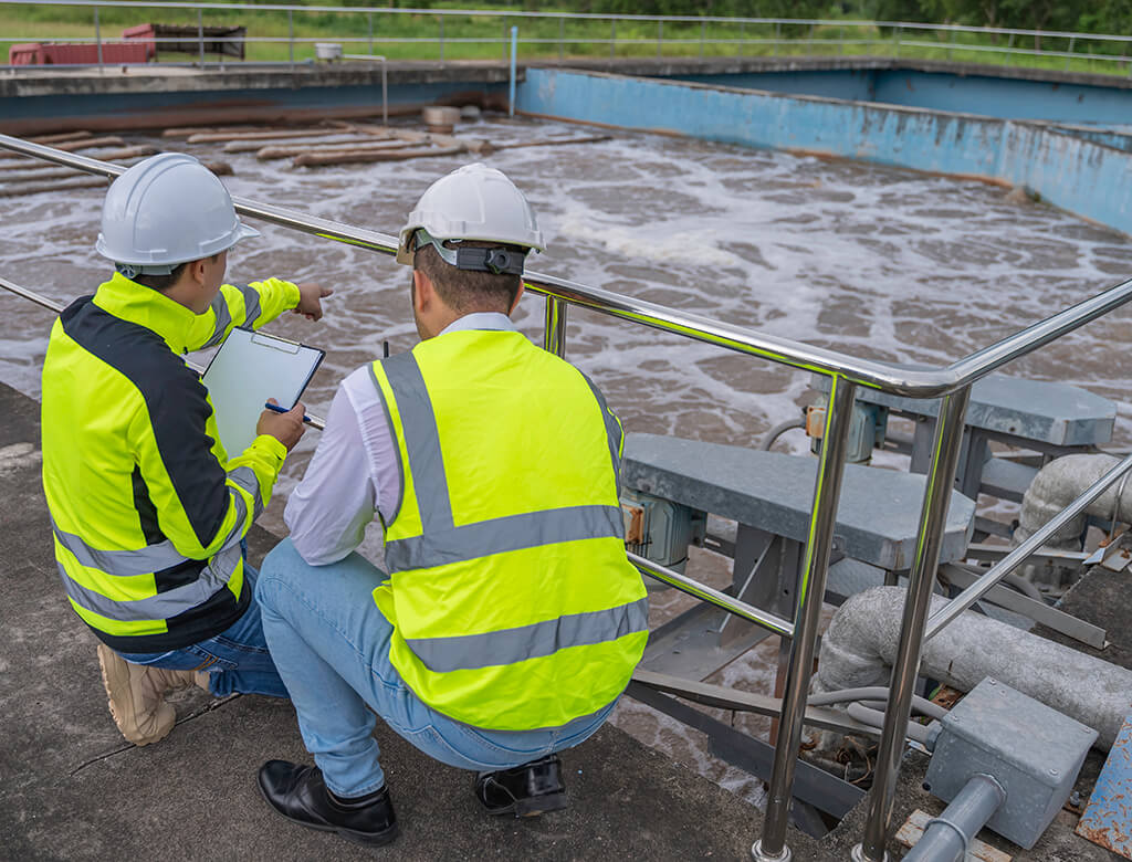 Wastewater engineers stoop by a stainless steel railing over a retention pond while discussing aeration piping. 